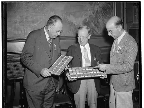 Three men in suits look over trays of old coins at a numismatic convention in Washington, D.C., in a black and white photograph