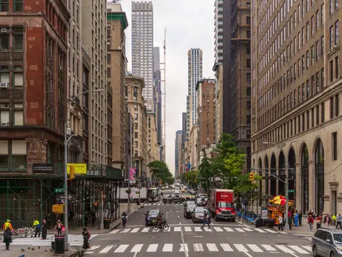 A Midtown Manhattan avenue on a bright day: stone office towers, shopfronts, a crosswalk and traffic running uptown