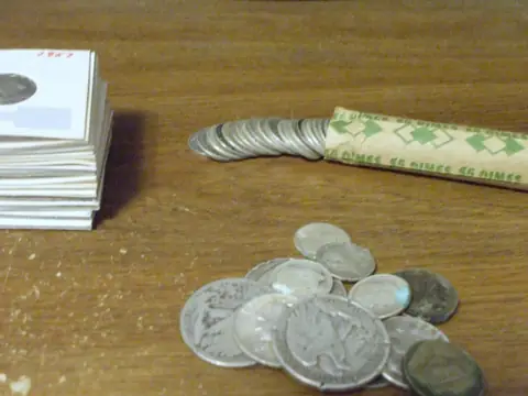 Junk silver on a wooden desk: silver dimes in paper coin holders, a roll of dimes and a loose pile of worn half dollars, quarters and dimes