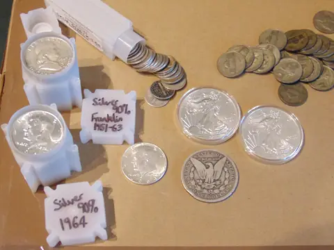 Investment silver on a sheet of card: tubes of Franklin and Kennedy half dollars, two Silver Eagles in capsules, a 1964 Kennedy half dollar, a worn Morgan dollar and a pile of wartime nickels